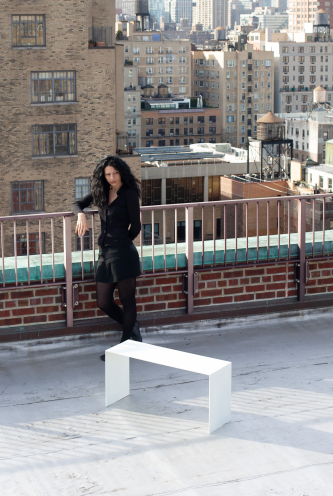 Lenka Ilic, a designer stands behind the bench on a roof top, resting on the parapet wall railing. NYC skyline in the background.